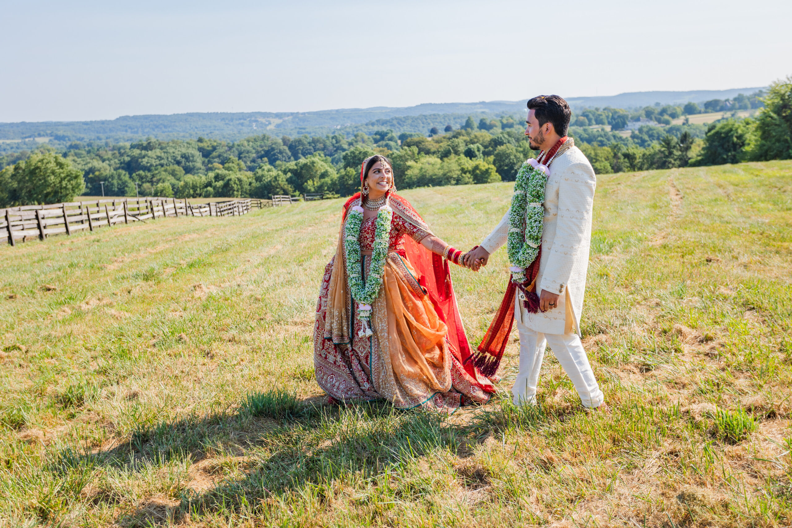 Bride & Groom dressed in traditional Indian garments hold hands and walk through an open field.