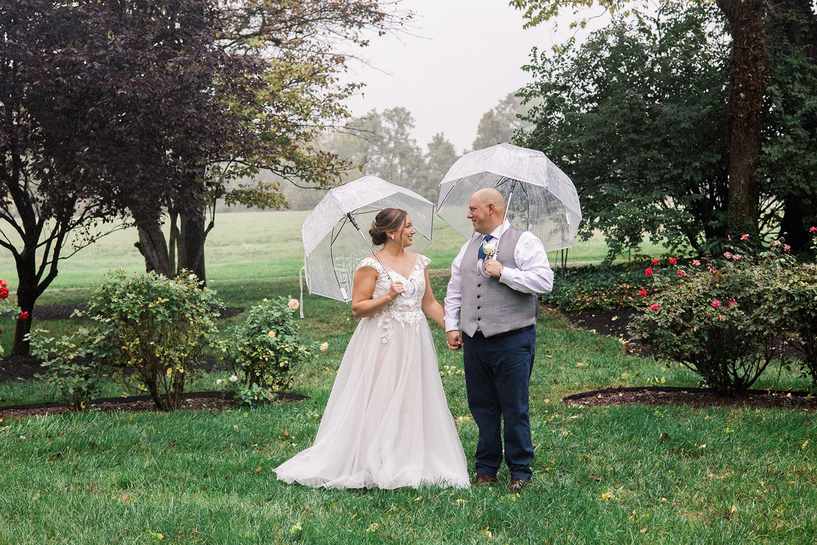 A Fredrick bride and groom share a romantic moment under a clear umbrella on their rainy wedding day, with soft lighting and lush greenery creating a magical atmosphere.