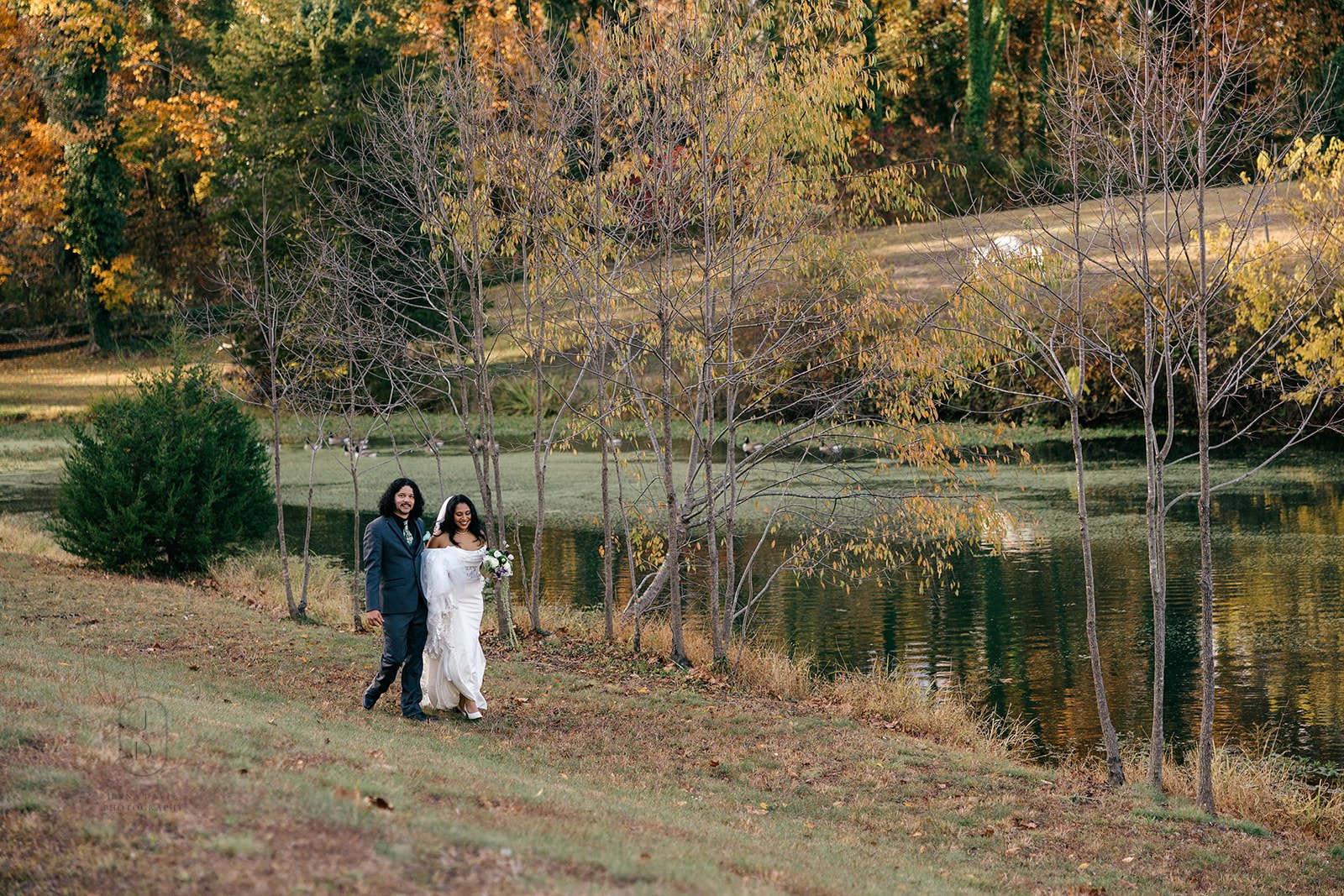 Bride and groom walking hand-in-hand beside pond at Poplar Springs Manor in fall